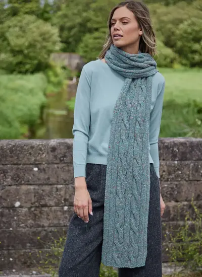 Woman styling a long sea foam Fisherman Donegal fleck cable scarf over a light blue sweater and grey trousers outdoors.